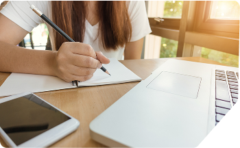 A student at a wooden desk using a laptop and taking notes, symbolising dissertation assistance by Scora for overwhelmed students.