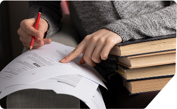 A person in a grey sweater holding a red pen and reviewing documents on a book pile, showcasing Scora’s expert help in academic proposal writing.